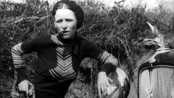 Bonnie Parker im Jahr 1933 steht mit Zigarre im Mund und Revolver in der Hand angelehnt an ihr Auto.
| Bild: picture alliance / Everett Collection | CSU Archives/Everett Collection