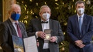 Reinhard Genzel (M), deutscher Physiker, hält bei der Übergabe des Nobelpreises für Physik die goldene Medaille in seinen Händen. Links steht dabei Per Thöresson, Schwedens Botschafter in Deutschland, mit der Urkunde, rechts steht Markus Söder (CSU), Ministerpräsident von Bayern. Wegen der Corona-Pandemie nehmen die Preisträger ihre Medaillen und Urkunden nicht in Stockholm, sondern an unterschiedlichen Orten entgegen. | Bild: picture alliance/dpa/dpa/Pool | Peter Kneffel