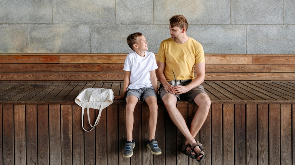 Vater und Sohn unterhalten sich, beide sitzen auf einer Holzbank. Was bedeutet Gewaltfreie Kommunikation mit Kindern? Der Psychologe Marshall B. Rosenberg hat sie entwickelt. Wir erklären, warum Gewaltfreie Kommunikation mit Kindern wichtig ist, wie ihr sie in vier Schritten erlernen könnt und was es mit der Giraffensprache auf sich hat.  | Bild: picture alliance/Westend61/Ekaterina Yakunina