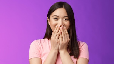 Junge Frau hält sich verlegen die Hände vors Gesicht. Schöne Komplimente bekommen wir alle gern, aber nicht allen fällt es leicht, sie anzunehmen - vor allem Frauen. Wie reagiert man richtig auf Komplimente, und was können sie bewirken? | Bild: picture alliance / Zoonar | Phongthorn Hiranlikhit