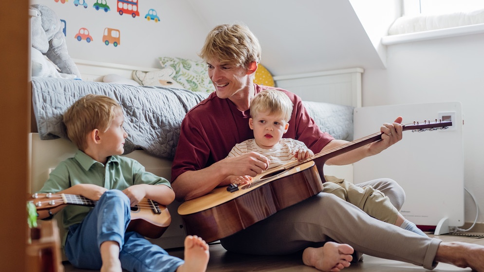 Ein Vater sitzt auf dem Boden mit seinen zwei Kindern und zupft an einer Gitarre. Signifikante Zeit verbringen mit den eigenen Kinder stärkt ihre soziale Kompetenz.  | Bild: colourbox.com