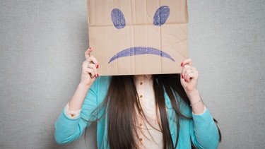 Frau mit Pappe vor dem Gesicht, die ein trauriges Smiley zeigt. Denkt ihr oft negativ? Wir erklären, warum manche Menschen eher Pessimisten oder Optimisten sind, was 'pessimistisch' bedeutet - und ob das wirklich immer schlecht ist. | Bild: colourbox.com