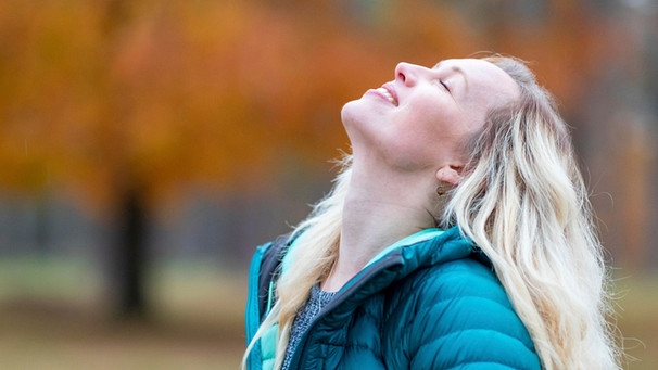 Eine Frau steht im Park und lächelt mit geschlossenen Augen. Selbstliebe bedeutet, mit sich selbst befreundet zu sein, zu seinen Bedürfnissen und Wünschen zu stehen und die eigenen Schwächen zu akzeptieren. Wie du lernst, dich selbst zu lieben. | Bild: picture alliance / Westend61 | William Perugini