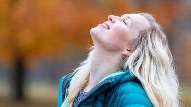 Eine Frau steht im Park und lächelt mit geschlossenen Augen. Selbstliebe bedeutet, mit sich selbst befreundet zu sein, zu seinen Bedürfnissen und Wünschen zu stehen und die eigenen Schwächen zu akzeptieren. Wie du lernst, dich selbst zu lieben. | Bild: picture alliance / Westend61 | William Perugini