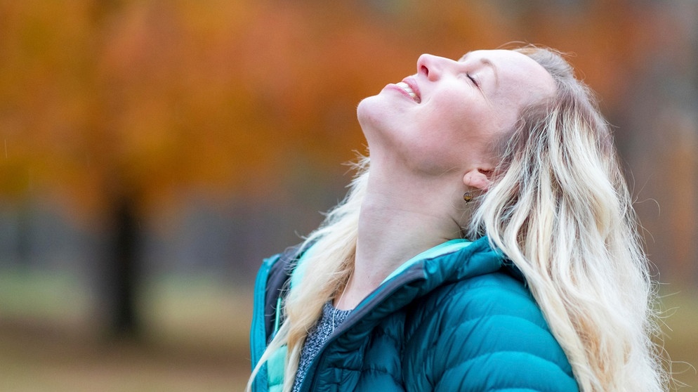 Eine Frau steht im Park und lächelt mit geschlossenen Augen. Selbstliebe bedeutet, mit sich selbst befreundet zu sein, zu seinen Bedürfnissen und Wünschen zu stehen und die eigenen Schwächen zu akzeptieren. Wie du lernst, dich selbst zu lieben. | Bild: picture alliance / Westend61 | William Perugini