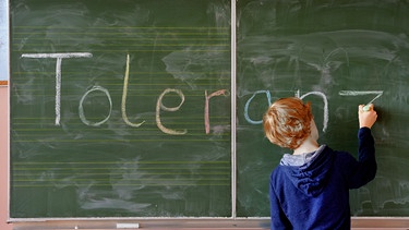 Schulkind schreibt Toleranz an die Tafel. Was für manche eine Zumutung ist, darüber zucken andere nur wohlwollend mit der Schulter. Woran liegt es, dass Menschen unterschiedlich tolerant sind? Und lässt sich Toleranz lernen? | Bild: picture alliance / Frank May | Frank May