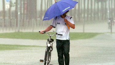 Ein Mann schiebt ein Fahrrad durch den Regen. Verbrennungen, Lähmungen, Bewusstlosigkeit, Herzstillstand: Ein Blitzschlag kann tödlich sein. Hier findet ihr Tipps zum richtigen Verhalten, wenn ihr unterwegs überrascht werdet. | Bild: picture-alliance/dpa